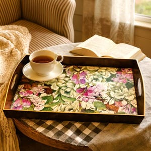 May include: A rectangular wooden serving tray with a floral design featuring pink and white flowers and green leaves. A white teacup and saucer sit on the tray, which has a dark brown border and gold trim. The tray is on a table with a checkered tablecloth.