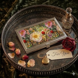 May include: A silver business card holder with a floral design featuring pink, yellow, and red flowers. The holder sits on a silver tray with a dried rose, a small perfume bottle, and a pendant with a tag that says "For You."