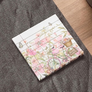May include: A square notepad with a floral and bicycle design. The notepad features a pink bicycle with a basket, flowers, and the text "She sells Seashells". The background has pink flowers and butterflies.