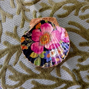 May include: A decorative scallop shell featuring a vibrant floral decoupage design on a black background. The shell has a prominent large pink flower at the center, surrounded by smaller orange, blue, and white blossoms, with a metallic gold painted edge.