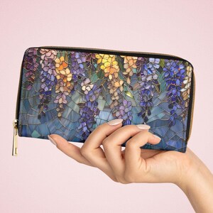 May include: A long, zippered wallet featuring a stained-glass design of wisteria flowers in shades of purple, yellow, and blue. The wallet has a black border and a gold-colored zipper and pull. The background is a mosaic of blue tiles.