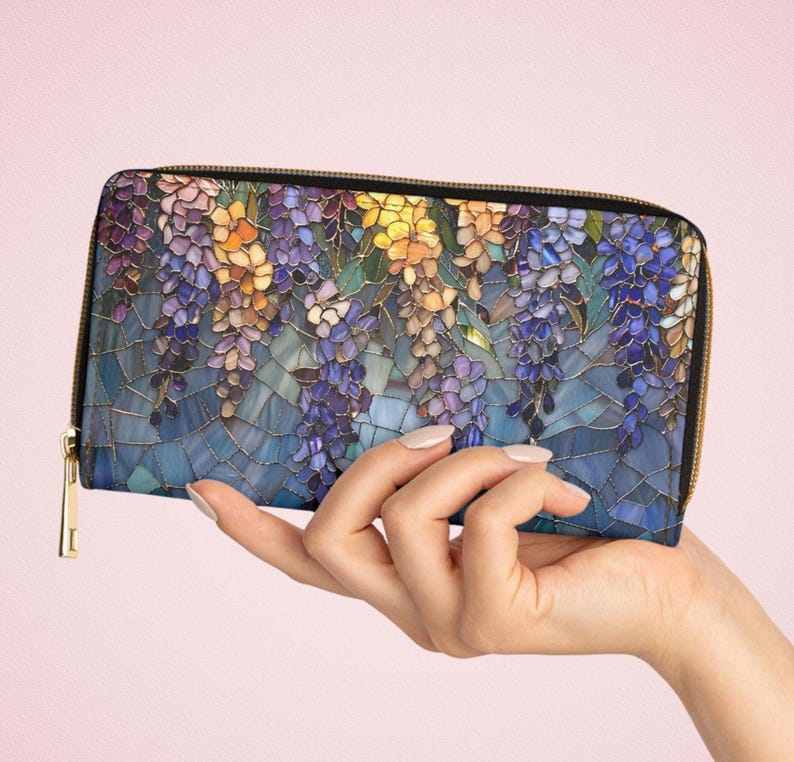 May include: A long, zippered wallet featuring a stained-glass design of wisteria flowers in shades of purple, yellow, and blue. The wallet has a black border and a gold-colored zipper and pull. The background is a mosaic of blue tiles.