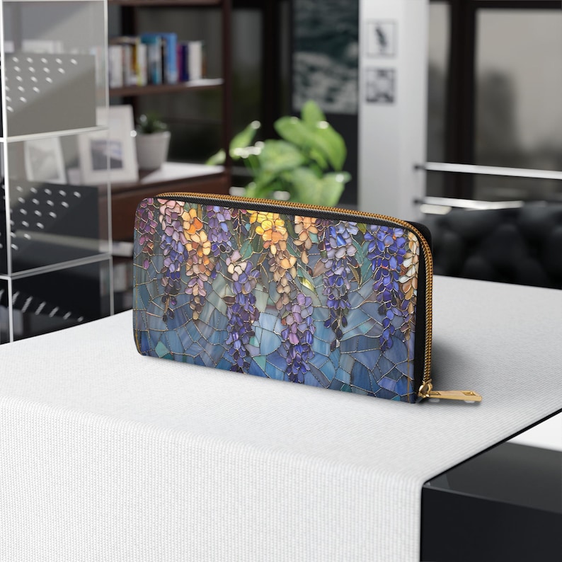 May include: A rectangular wallet with a stained glass design featuring purple, yellow, and blue wisteria flowers. The wallet has a gold zipper and a black border. The design is reminiscent of a mosaic.