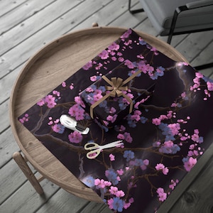 Può includere: Un regalo avvolto in carta da regalo floreale nera con fiori rosa e blu e un nastro dorato. Un rotolo di nastro bianco e forbici dorate sono sul tavolo. Il tavolo è di legno.