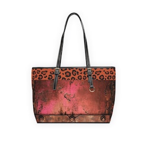 May include: A tote bag with black handles and straps. The bag features a rusty, textured design with a pink center and a leopard print band at the top. The bag has a rectangular shape and is made of a durable material.