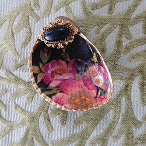 May include: A decorative oyster shell dish featuring a black interior with a vibrant floral print of pink and yellow flowers. The rim is painted with a metallic gold finish, and an oval black cabochon set in an ornate gold frame is attached.