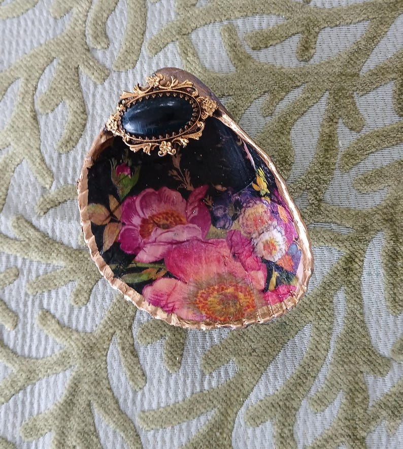 May include: A decorative oyster shell dish featuring a black interior with a vibrant floral print of pink and yellow flowers. The rim is painted with a metallic gold finish, and an oval black cabochon set in an ornate gold frame is attached.
