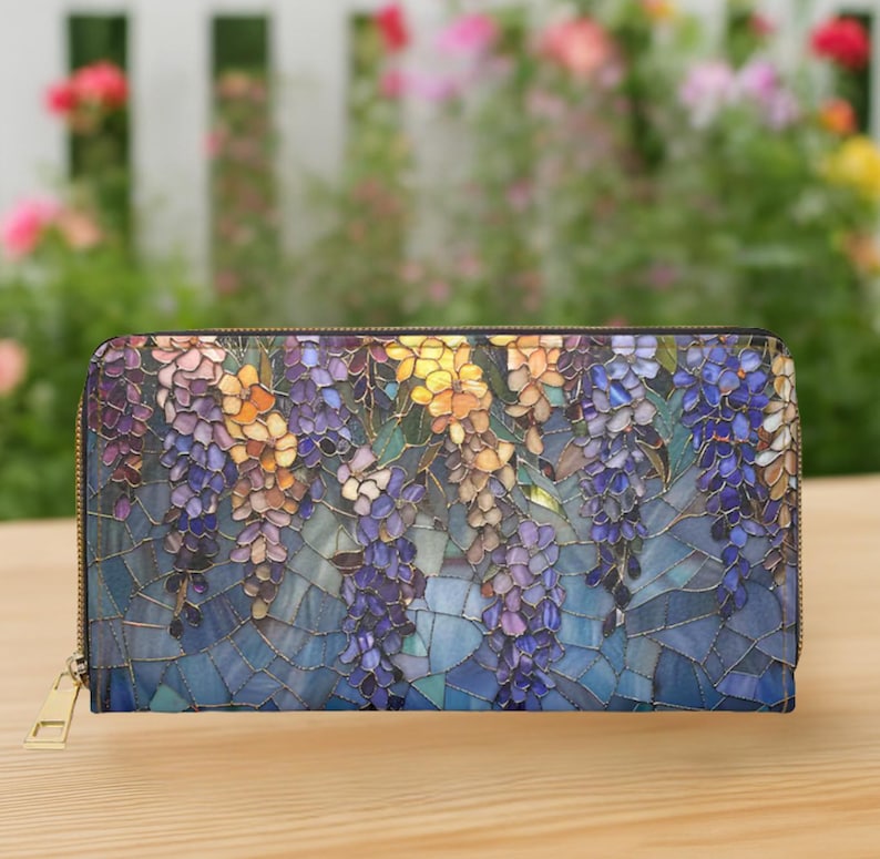 May include: A zippered wallet featuring a mosaic-style floral design with purple, yellow, and blue wisteria blooms. The wallet has a gold-colored zipper and a rectangular shape. The background is blurred, with a wooden surface.