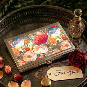 May include: A rectangular metal case featuring a colorful floral stained glass pattern on the lid. The case sits on a dark tray alongside a small glass perfume bottle, a dark red rose, scattered rose petals, and a gold tag reading For You.