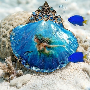 Puede incluir: Un adorno de concha decorativo que presenta una escena submarina de color azul vibrante con una ilustración de sirena. La concha está rematada con un adorno de filigrana de metal color bronce y decorada con pequeñas cuentas azules.