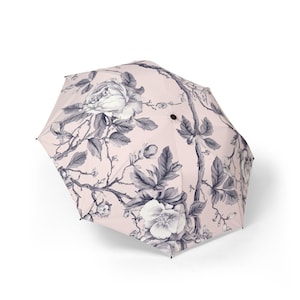 Può includere: Un ombrello pieghevole con una copertura rosa chiaro decorata con una stampa botanica grigio scuro di fiori in fiore, rami frondosi e boccioli. L'ombrello presenta un perno centrale nero e punte nere alle estremità delle stecche metalliche.