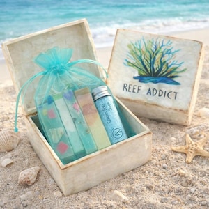 May include: A square white box with a lid featuring a blue and green coral illustration and the text REEF ADDICT. Inside, a turquoise organza bag holds four rectangular soap bars and a small glass bottle labeled Epsom Salt Soak.