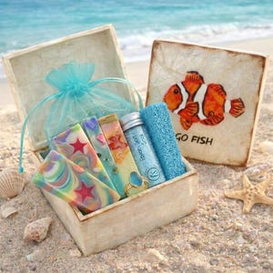 May include: A gift box with a lid featuring a clownfish illustration and the text GO FISH, containing colorful swirl-patterned soap bars with star accents, a small bottle of Epsom salt soak, a blue textured sponge, and a gold-toned ring.