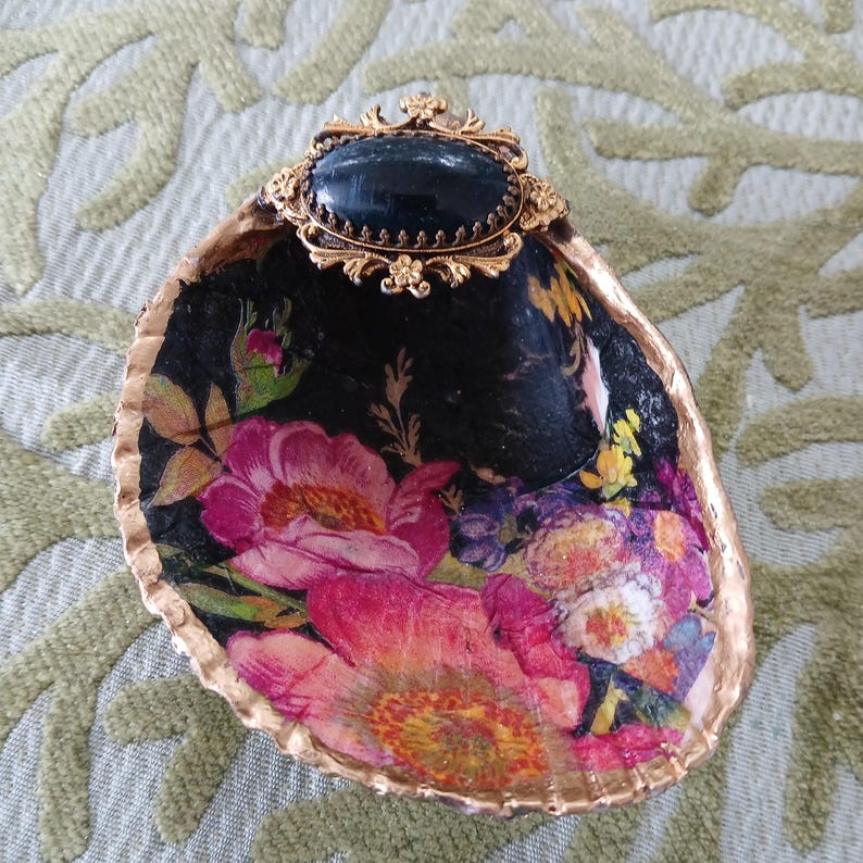 May include: A decorative oyster shell featuring a decoupage floral pattern with vibrant pink and yellow flowers on a black background. The shell edge is painted gold, and an oval black cabochon set in an ornate gold-tone metal frame is attached.