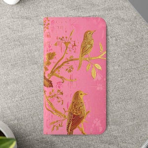 May include: A rectangular pink wallet featuring a gold foil print of two birds perched on branches with leaves and flowers. The design covers the front surface, showcasing a metallic gold finish against a solid pink background with a textured appearance.