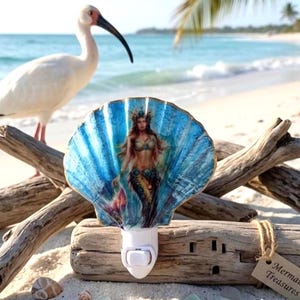 May include: A decorative night light featuring a large, fan-shaped seashell painted in shades of blue. The shell displays a detailed illustration of a mermaid with long hair and a tail. The light is mounted on a white plastic plug-in base.