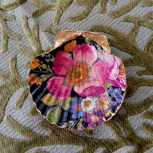 May include: A decorative scallop shell featuring a vibrant floral decoupage design with large pink flowers, smaller blue and yellow blossoms, and green leaves set against a black background, finished with a metallic copper-colored gilded edge.