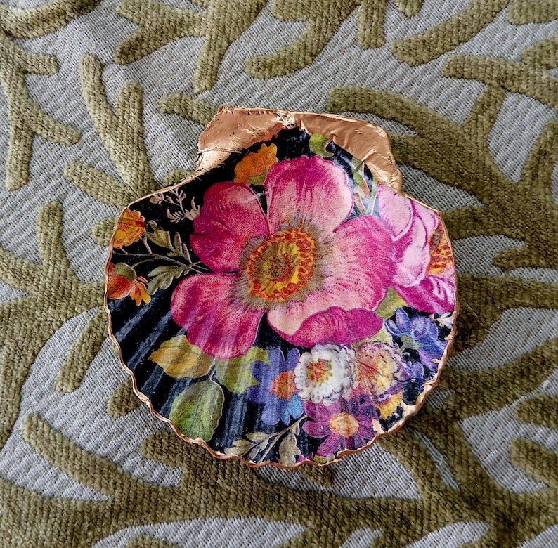 May include: A decorative scallop shell featuring a vibrant floral decoupage design with large pink flowers, smaller blue and yellow blossoms, and green leaves set against a black background, finished with a metallic copper-colored gilded edge.