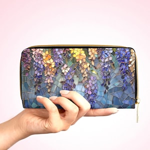 May include: A zippered wallet featuring a mosaic-style floral design with purple, yellow, and blue wisteria blossoms. The wallet has a gold-colored zipper and is held against a light pink background.