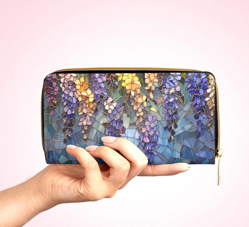 May include: A zippered wallet featuring a mosaic-style floral design with purple, yellow, and blue wisteria blossoms. The wallet has a gold-colored zipper and is held against a light pink background.