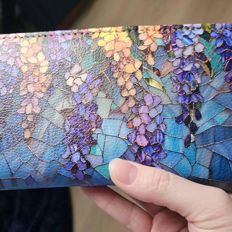 May include: A colorful wallet with a floral design. The wallet features a mosaic-like pattern of purple, blue, yellow, and orange flowers and leaves. The background is a gradient of blues. The wallet is held by a hand.