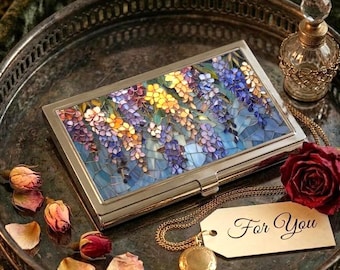 Op de afbeelding: Een rechthoekig zilverkleurig metalen doosje met een mozaïek van blauwe, paarse en gele blauweregenbloemen. Het wordt vergezeld door een goudkleurig medaillon aan een ketting met een label met de tekst For You, een glazen parfumflesje en rozenblaadjes.
