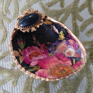 May include: A decorative seashell dish featuring a decoupage floral pattern with vibrant pink, yellow, and purple flowers on a black background. The shell edge is painted gold, and a black oval cabochon in a gold filigree setting is attached.