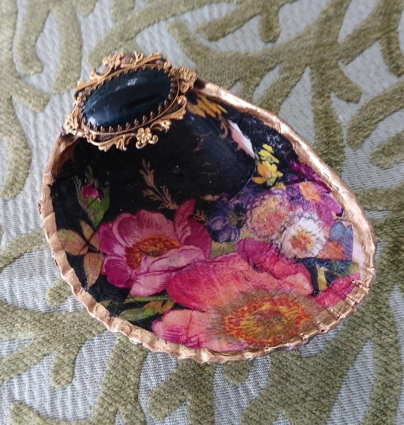 May include: A decorative seashell dish featuring a decoupage floral pattern with vibrant pink, yellow, and purple flowers on a black background. The shell edge is painted gold, and a black oval cabochon in a gold filigree setting is attached.