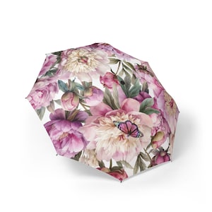 Peut inclure: Un parapluie pliable présentant un fond blanc avec un motif dense de pivoines en fleurs roses et crème, un feuillage vert et un papillon aux ailes roses et noires posé sur l'un des grands pétales de la fleur.