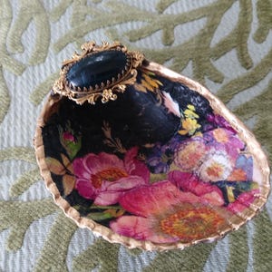 May include: A decorative seashell dish featuring a decoupage floral pattern with pink, red, and yellow flowers on a black background. The shell edge is painted gold, and a gold-toned filigree brooch with a black oval stone is attached to the top.