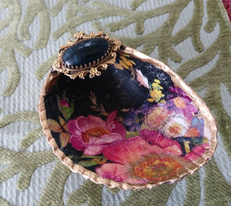May include: A decorative seashell dish featuring a decoupage floral pattern with pink, red, and yellow flowers on a black background. The shell edge is painted gold, and a gold-toned filigree brooch with a black oval stone is attached to the top.