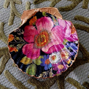 May include: A decorative scallop shell featuring a vibrant floral print on a black background. The design displays large pink, orange, blue, and white flowers with green leaves. The scalloped edges of the shell are finished with a metallic copper trim.