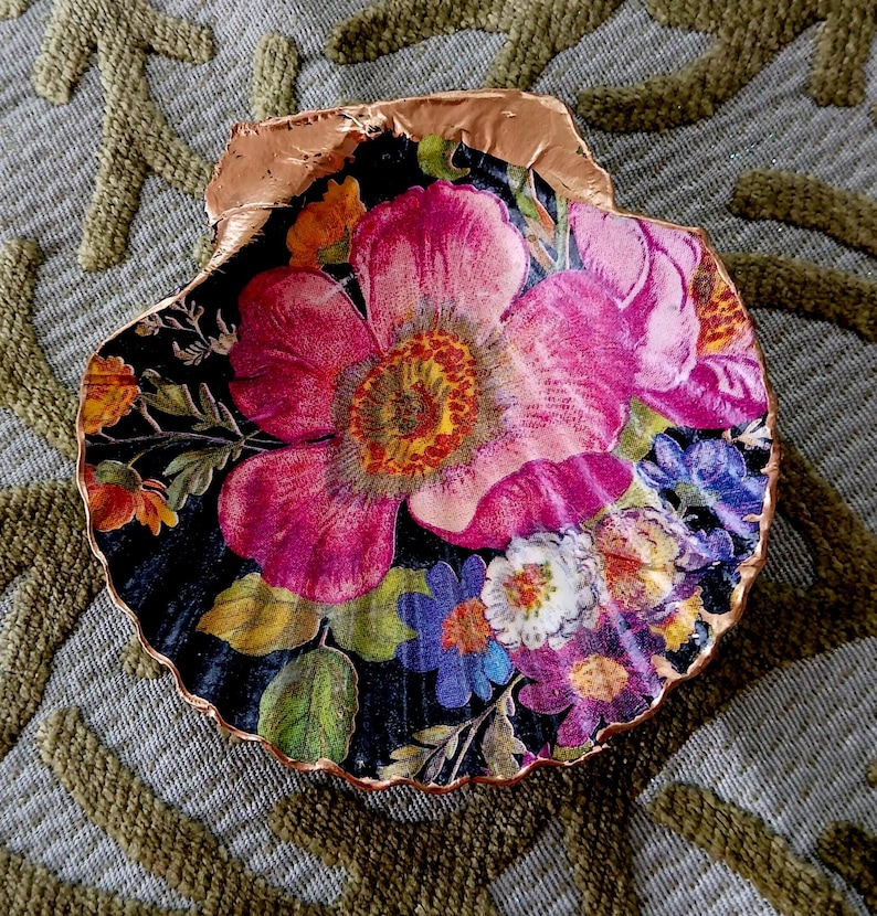 May include: A decorative scallop shell featuring a vibrant floral print on a black background. The design displays large pink, orange, blue, and white flowers with green leaves. The scalloped edges of the shell are finished with a metallic copper trim.