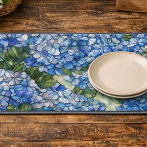 Puede incluir: Un tapete rectangular para secar platos con un diseño floral de hortensias en tonos azules y verdes. Dos platos beige apilados sobre el tapete. El tapete tiene un borde gris.