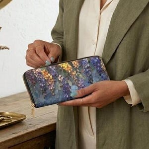 May include: A long, zippered wallet with a floral design in shades of blue, purple, yellow, and white. The wallet has a black border and a gold-colored zipper pull. The wallet is held by a person wearing a green jacket.