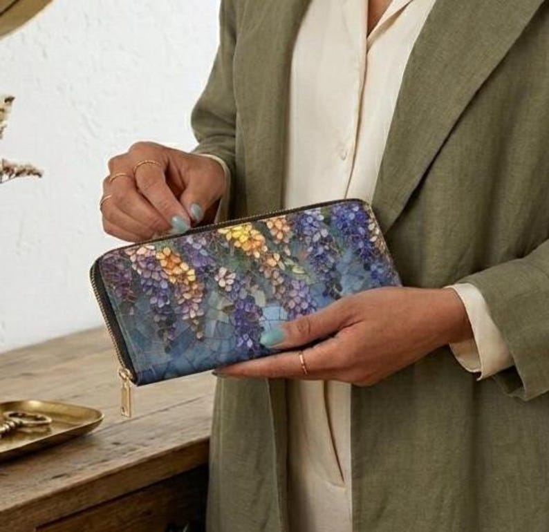 May include: A long, zippered wallet with a floral design in shades of blue, purple, yellow, and white. The wallet has a black border and a gold-colored zipper pull. The wallet is held by a person wearing a green jacket.