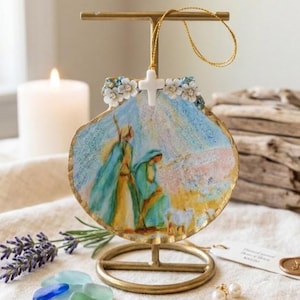 May include: A scallop shell ornament featuring a painted nativity scene with figures in blue and gold robes. The shell is topped with a small white cross and white floral accents, suspended by a gold cord on a gold metal display stand.