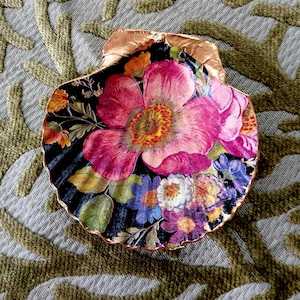 May include: A decorative scallop shell featuring a vibrant floral decoupage design with large pink, purple, and yellow flowers on a black background. The scalloped edges of the shell are finished with a metallic copper-colored trim.