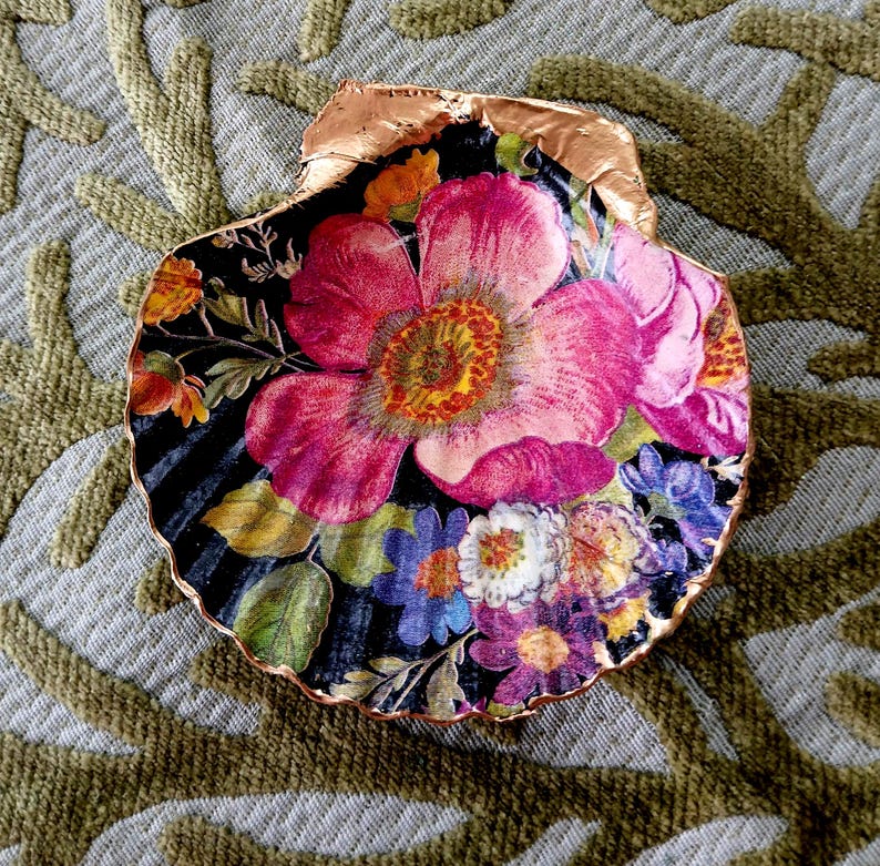 May include: A decorative scallop shell featuring a vibrant floral decoupage design with large pink, purple, and yellow flowers on a black background. The scalloped edges of the shell are finished with a metallic copper-colored trim.