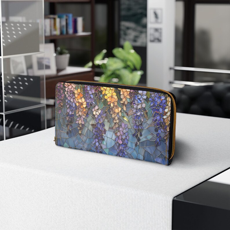 May include: A zippered wallet featuring a stained-glass style floral design in shades of purple, yellow, and blue. The wallet has a gold-colored zipper and a black border. The design depicts hanging wisteria flowers.