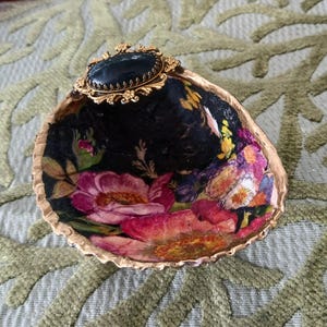 May include: A decorative seashell dish featuring a black interior with a vibrant floral pattern of pink and yellow flowers. The rim is painted gold, and a gold-toned oval brooch with a dark blue cabochon is attached to the top edge of the shell.