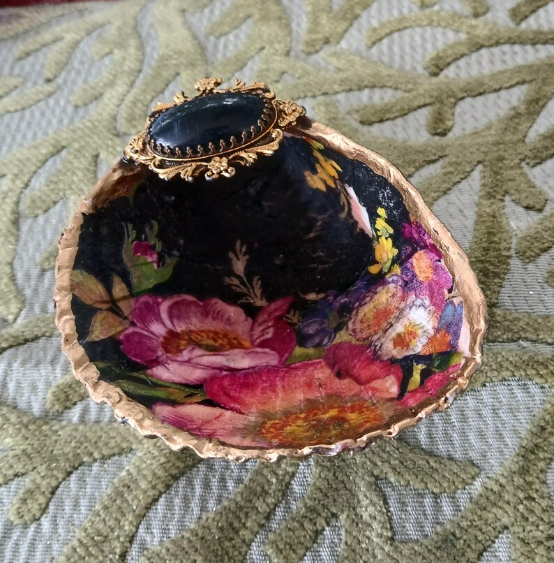 May include: A decorative seashell dish featuring a black interior with a vibrant floral pattern of pink and yellow flowers. The rim is painted gold, and a gold-toned oval brooch with a dark blue cabochon is attached to the top edge of the shell.