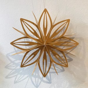 May include: A gold paper star ornament featuring multiple layered, pointed petals with cut-out centres. The Christmas star is suspended by a thin string and displays a symmetrical, multi-dimensional design with sharp edges and a matte finish.