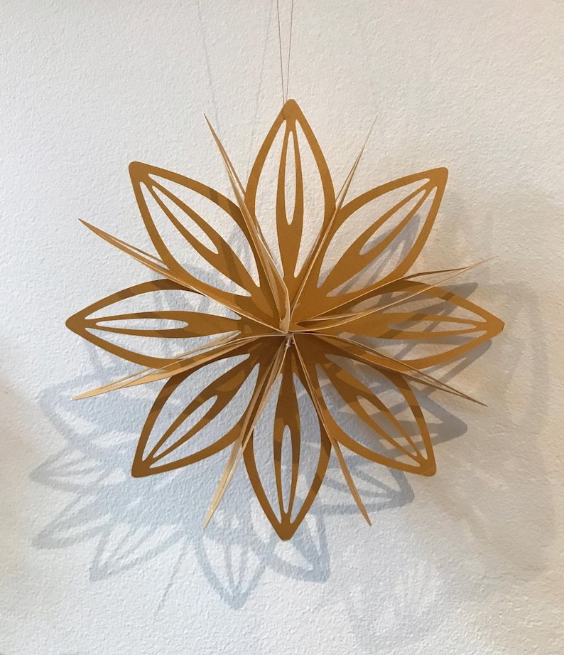 May include: A gold paper star ornament featuring multiple layered, pointed petals with cut-out centres. The Christmas star is suspended by a thin string and displays a symmetrical, multi-dimensional design with sharp edges and a matte finish.