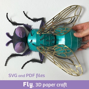 May include: A 3D paper craft fly featuring a metallic teal body, purple dotted eyes, and intricate gold-toned translucent wings. The insect has black legs and antennae. Text at the bottom reads: SVG and PDF files, Fly, 3D paper craft.