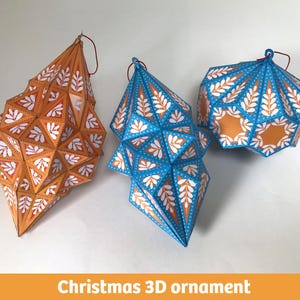 Puede incluir: Tres adornos geométricos en 3D con estructura facetada. Uno es naranja con patrones de hojas blancas, otro es azul con patrones de hojas blancas y el tercero es una forma más corta azul y naranja con patrones de hojas blancas. Cada uno tiene un lazo rojo.