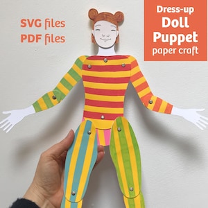 May include: A paper craft dress-up doll puppet with articulated limbs connected by metal fasteners. The doll features brown hair in buns, a yellow and red striped torso, green and yellow striped arms, and blue and yellow striped legs. Text reads: SVG files, PDF files, Dress-up Doll Puppet paper craft.