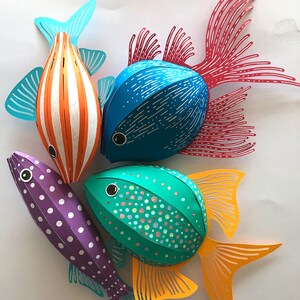 May include: Four three-dimensional paper fish decorations. One is orange with white stripes and teal fins, one is blue with white dashes and red fins, one is purple with white polka dots, and one is teal with multicolored dots and yellow fins.