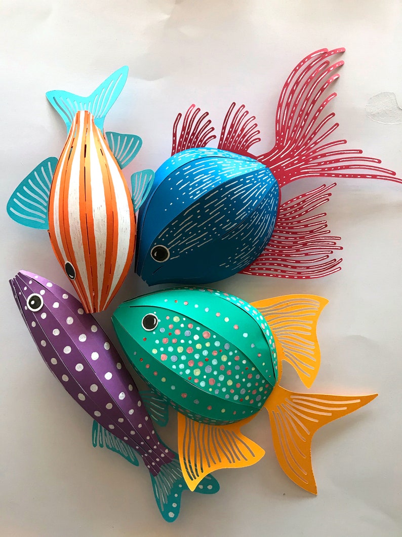 May include: Four three-dimensional paper fish decorations. One is orange with white stripes and teal fins, one is blue with white dashes and red fins, one is purple with white polka dots, and one is teal with multicolored dots and yellow fins.