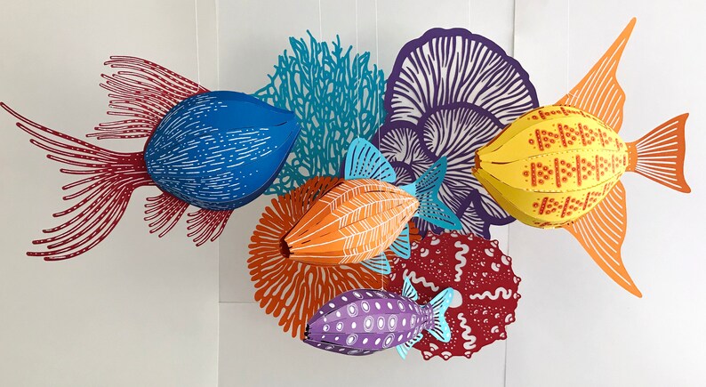 May include: A hanging mobile featuring three-dimensional paper fish in blue, orange, and purple, accompanied by flat, intricate paper cutouts of coral, sea anemones, and sea urchins in shades of red, orange, blue, and purple against a white background.
